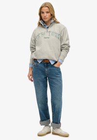 Sweat-shirt gris avec le texte "NEW YORK", superposé sur une chemise bleu clair. Porté avec un jean bleu ample, aux manches retroussées, et des baskets de couleur crème.