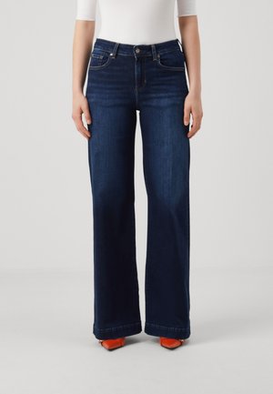 Persona che indossa jeans blu scuro a gamba larga e scarpe appuntite arancioni brillanti, in piedi contro uno sfondo chiaro uniforme.