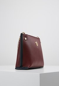 Bolso de piel burdeos con acentos en azul marino, que presenta un logo en tono dorado y una correa de cadena. Forma estructurada y superficie texturizada.