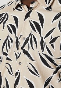 Camisa de manga corta en tejido beige con un estampado de hojas en blanco y negro, con cierre de botones al frente y cuello clásico.