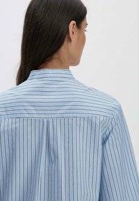 Camicia a righe blu chiaro con collo alto, piega sul retro e maniche lunghe. Sottili righe scure corrono verticalmente su uno sfondo blu solido.