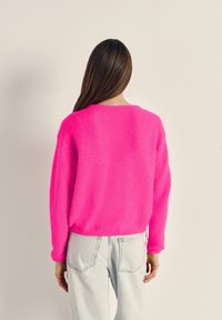 Moteris su ilgais rudos spalvos plaukais, dėvinti ryškiai rožinį megztinį ir šviesiai mėlynas džinsus, stovinti nugara priešais vienspalvį smėlio spalvos foną.