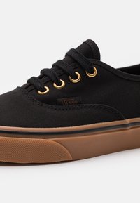 Sneaker nera in tela con punta rotonda, lacci neri, occhielli dorati e suola in gomma marrone. Presenta un logo Vans sul lato.