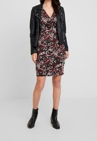 Kvinna iklädd en svart skinnjacka över en röd, vit och svart paisleymönstrad klänning med svarta ankelstövlar, stående mot en enkel bakgrund.