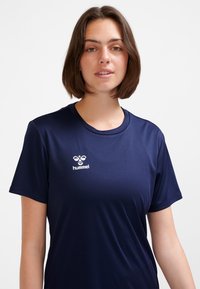 Ung kvinne med skulderlangt brunt hår iført en marineblå kortermet sportsskjorte med hvit Hummel-logo på brystet.