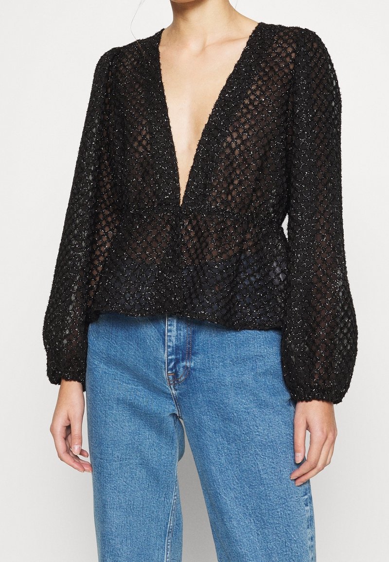 Blouse noire à manches longues avec décolleté en V profond, confectionnée en tissu en maille texturée, dotée d'un ourlet en peplum et d'un subtil scintillement. Assortie à un jean bleu.