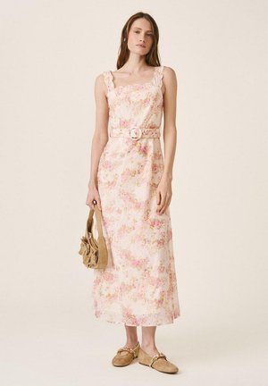 Mujer con un vestido largo floral sin mangas y cinturón, sosteniendo un bolso marrón y usando zapatos planos decorativos beige sobre un fondo liso.