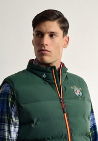 Chaleco acolchado verde con cuello alto, cremallera frontal y logo bordado. Debajo, se ve una camisa a cuadros azul y amarilla.