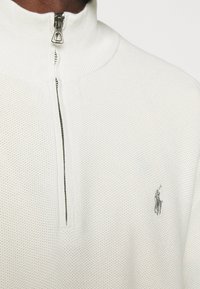 Weißer Strickpullover mit hohem Kragen und silbernem Reißverschluss, ausgestattet mit einem kleinen gestickten Logo auf der linken Seite. Die Textur ist fein und atmungsaktiv.