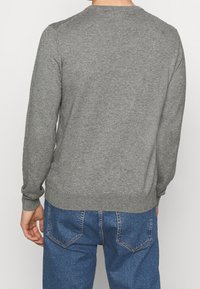 Mann in einem schlichten grauen Langarmpullover und blauen Jeans, der mit dem Rücken zur Kamera vor einem einfarbigen Hintergrund steht.