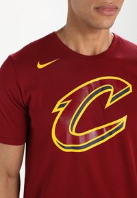 Camiseta de algodón color marrón oscuro con un gran logo "C" en amarillo y azul marino en el frente y un pequeño swoosh de Nike en amarillo en la parte superior izquierda. Cuello redondo.
