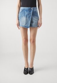 Falda envolvente de mezclilla en azul claro, con cierre de botones, dos bolsillos laterales y dobladillo deshilachado. Combinada con mule de tacón negro.