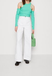 Nainen pukeutuneena turkoosiin kylmät olkapäät paljastavaan pitkähihaiseen puseroon, valkoisiin korkeavyötäröisiin leveälahkeisiin housuihin, mustiin kenkiin ja pitää vihreää käsilaukku.