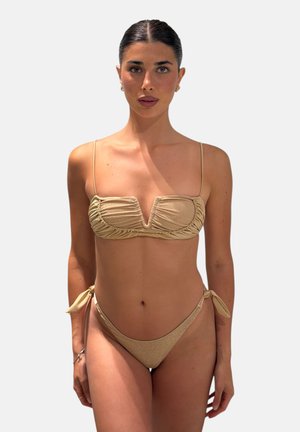 Donna con capelli scuri in bikini dorato con top arricciato e slip con lacci laterali, in piedi contro uno sfondo chiaro e uniforme.