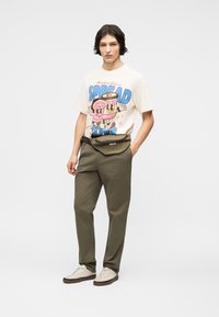 Νέος με σκούρα μαλλιά φοράει λευκό T-shirt με γραφικό σχέδιο, ελιάς πράσινο παντελόνι, μπεζ αθλητικά παπούτσια και μια τσάντα μέσης σε τόνους ελιάς πράσινου.