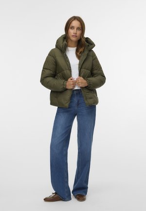 Junge Frau trägt eine olivgrüne Steppjacke mit Kapuze, weißes T-Shirt, blaue Schlagjeans und braune Schuhe, steht vor weißem Hintergrund.