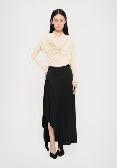 Beige top met cowl-hals en lange mouwen, gecombineerd met een zwarte asymmetrische lange rok met een gladde textuur en een aansluitende silhouet. Zwarte puntige hakken.