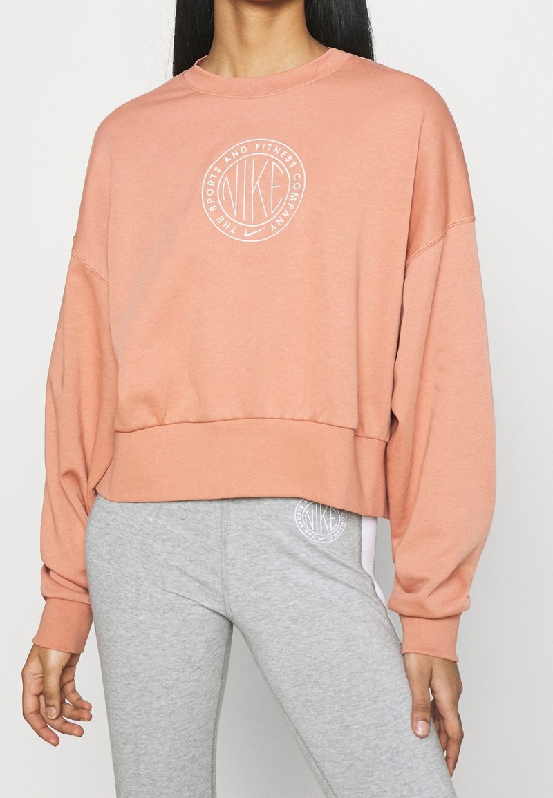 Klädd i en kortärmad sweatshirt i mjuk persika med överdimensionerade ärmar, som har en vit cirkulär Nike-logotyp i mitten. Matchad med grå leggings.