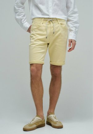 Hombre con pantalones cortos enrollados de color amarillo pálido con cordón blanco, camisa blanca de manga larga y zapatillas casuales beige, de pie contra un fondo liso.