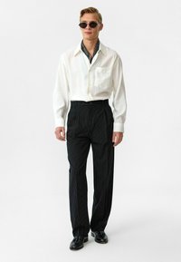 Witte langemouwen shirt met een borstzak, gecombineerd met zwarte streepjasken, en zwarte schoenen; geaccessoireerd met ronde zonnebrillen.