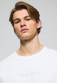 Biała bawełniana koszulka z okrągłym dekoltem oraz wytłoczonym logo "Polo Club" z przodu, gładka tekstura i klasyczny krój.