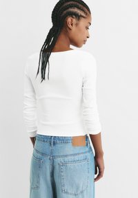 Haut à manches longues blanc à nervures avec un décolleté carré, associé à un pantalon en denim large bleu clair avec une patch en cuir brun au dos.