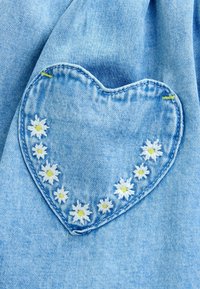 Tissu en denim bleu clair avec un patch en forme de cœur. Le patch présente des marguerites blanches aux centres jaunes, entourées de détails de couture bleue.