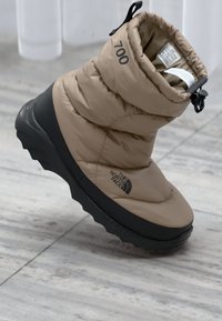 Botte isolée beige avec un design épais, semelle en caoutchouc noire et détail de logo. Dispose d'un cordon de serrage en haut pour un ajustement.