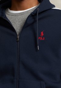 Marinblå zip-up hoodie gjord av texturerat tyg, med en röd broderad logotyp, silverfärgad dragkedja och en grå crew neck under.