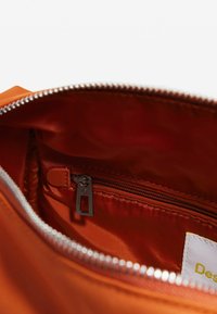 Interno di una borsa arancione con texture satinata liscia, dotato di una tasca con zip e hardware della cerniera in argento. Etichetta con "Design" visibile.