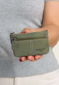 Ruka drží malou zelenou koženou mincovnici na zip s bílým prošíváním a logem "Gusti Leder studio".