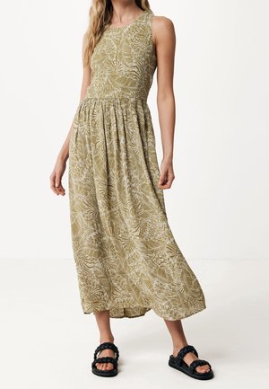 Robe maxi en vert olive avec un motif de feuilles, sans manches, taille froncée et jupe fluide. Associée à des sandales noires à lanières.