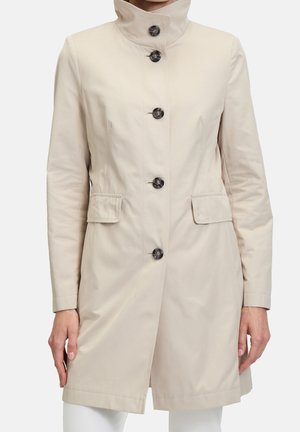 Manteau beige longueur genou avec col haut, manches longues, cinq gros boutons et poches avant à rabat porté par une personne debout.