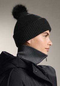 Czarna prążkowana dzianinowa czapka z pomponem z futra, zestawiona z szarym swetrem z wysokim kołnierzem i czarną puchową kurtką. Widok z profilu.