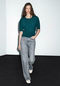 Türkisfarbenes Kurzarm-Stricktop kombiniert mit hochgeschnittenen, hellgrauen, geraden Jeans und weißen Slippern vor einem minimalistischen Hintergrund.