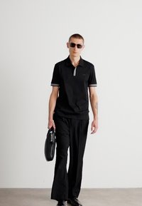 Camisa polo preta com detalhes em cinza no colarinho, combinada com calças pretas e bolsa de couro. Modelo usando óculos de sol, em pé sobre uma superfície lisa.