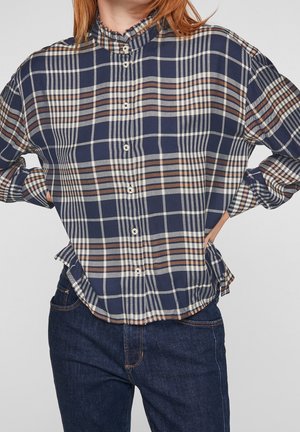 Kvinde iført en ternet skjorte i marineblå, brun og hvid med flæsekrave og mørkeblå jeans, hænderne på hofterne.