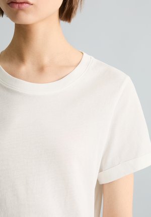 Młoda osoba nosząca gładką białą koszulkę z krótkim rękawem i okrągłym dekoltem, widoczna od szyi do górnej części piersi i prawego ramienia na jasnym tle.