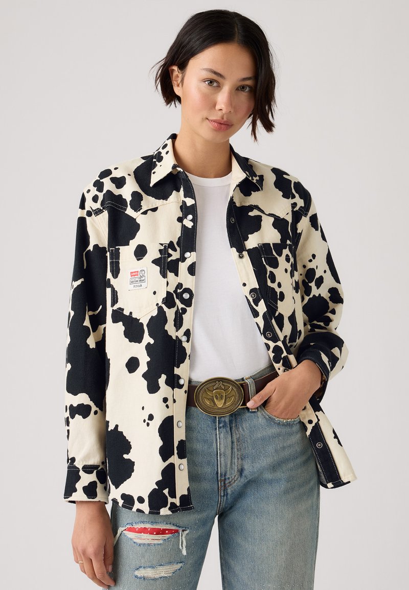 Svart och krämfärgad kappa med cow-print, spetsig krage, framfickor och metallknappar; parat med ljusblå slitna jeans och ett dekorativt bälte.
