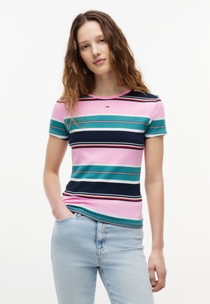 Νεαρή γυναίκα με κυματιστά καστανά μαλλιά φοράει ένα ριγέ κοντομάνικο τοπ σε ροζ, τιρκουάζ και ναυτικό μπλε χρώμα και ανοιχτόχρωμα τζιν, στέκεται με το ένα χέρι στην τσέπη.