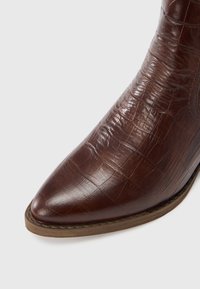 Brązowy botek na kostkę ze sztucznej skóry, z wydłużonym noskiem, teksturowanym wzorem krokodyla oraz subtelnymi przeszyciami wzdłuż cholewki i podeszwy.