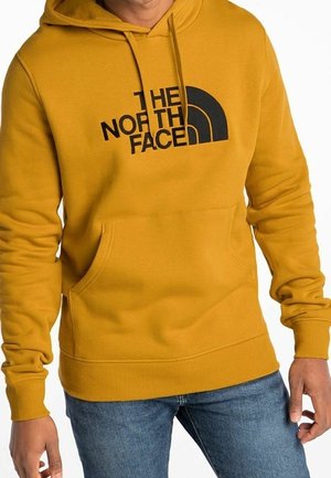 Mężczyzna w musztardowo-żółtej bluzie z czarnym logo "The North Face" i niebieskich dżinsach, stoi z rękami swobodnie opuszczonymi wzdłuż ciała.