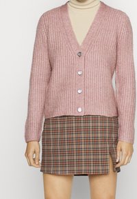 Rosa stickad kofta med V-hals, fem silverknappar och ribbad textur. Kombinerad med en brun rutmönstrad mini kjol med en sidoöppning.