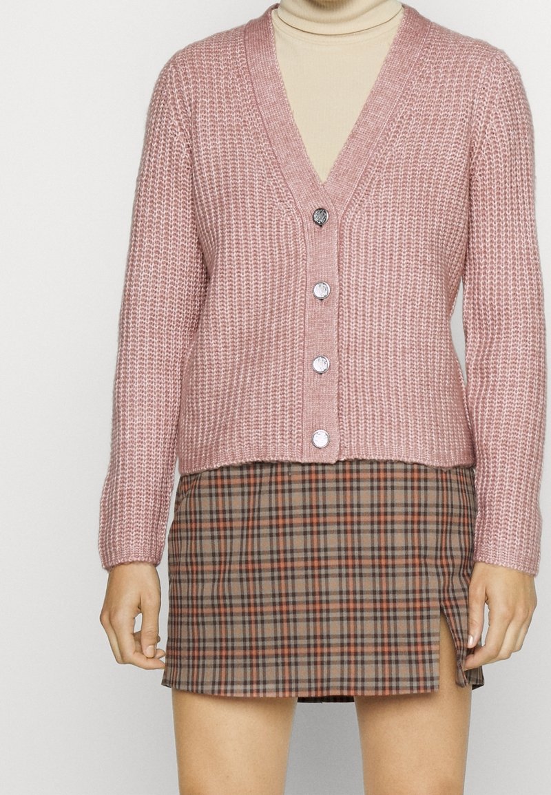 Rosa stickad kofta med V-hals, fem silverknappar och ribbad textur. Kombinerad med en brun rutmönstrad mini kjol med en sidoöppning.