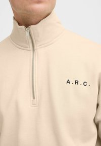 Beige ritssluiting fleece pullover met een hoge kraag, voorzien van een zwart "A.R.C." logo op de linkerborst. Gladde textuur, middelzwaar materiaal.