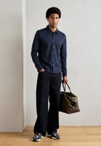 Marineblå knapp-up skjorte, mørke vide bukser og flerfargede joggesko. Holder en olivengrønn duffelbag med brune detaljer.