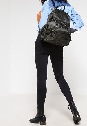 Person trägt schwarze Skinny-Jeans, schwarze Stiefeletten und eine blaue Jeansjacke mit einem grünen Rucksack im Tarnmuster, steht vor einem weißen Hintergrund.