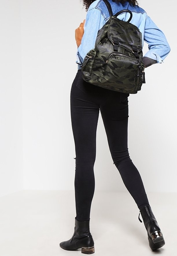 Personne portant un jean skinny noir, des bottines noires et une veste en jean bleue avec un sac à dos camouflage vert, debout devant un fond blanc.