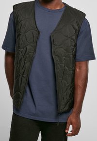 Černá prošívaná vesta se zipem vpředu, zdobená vlnitými vzory, nošená přes námořnické modré tričko. K dispozici v různých velikostech a střizích.