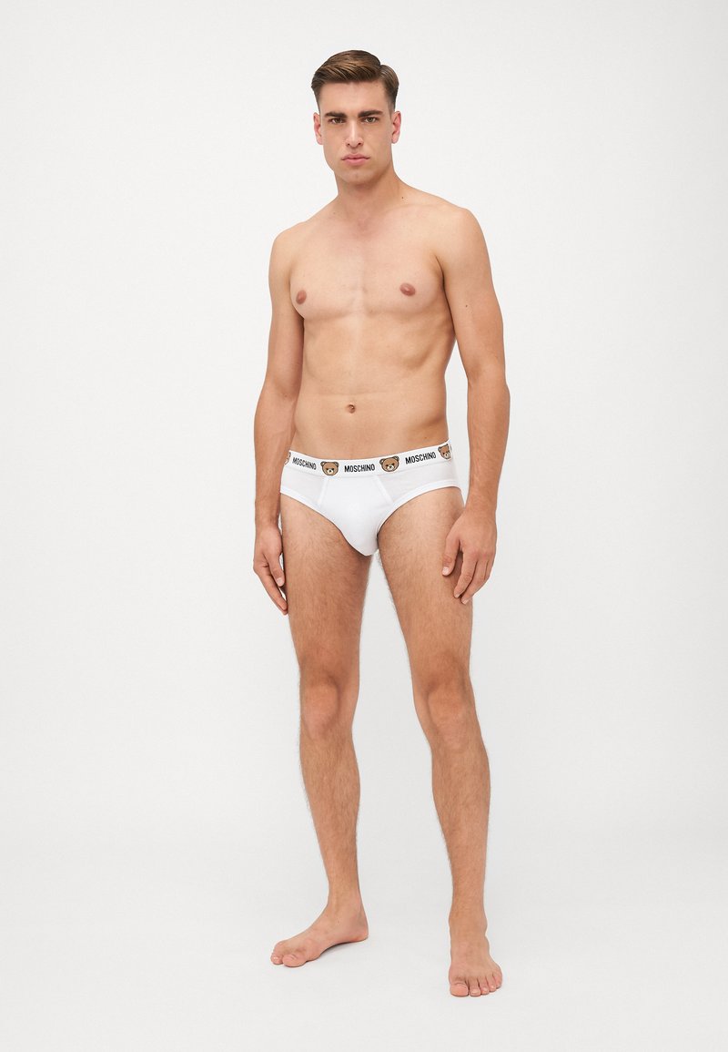 Weiße Baumwollslips mit schwarzem Bund, der das "MOSCHINO"-Logo und eine Bären-Grafik zeigt. Modell steht barfuß, Hände an den Seiten, Frontalansicht.